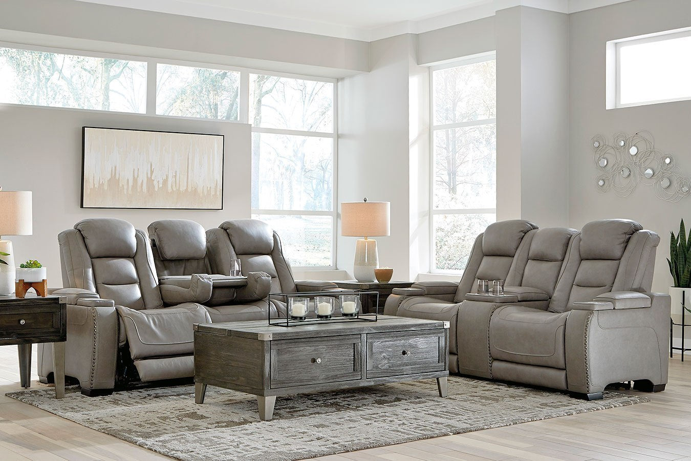 A modern living room features two gray leather reclining sofas, a wooden coffee table with storage drawers, and two side tables with lamps. The space is adorned with a wall painting, decorative wall art, and large windows that let in natural light.