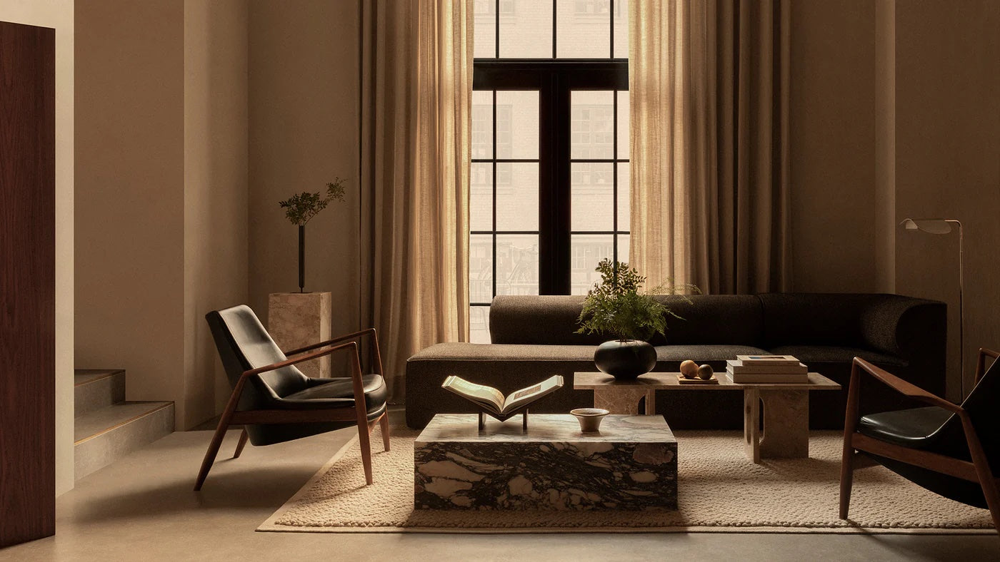 A cozy, modern living room featuring a large window with beige curtains, a dark sofa, two black armchairs, and a low marble coffee table. The table holds an open book, a vase with flowers, and books. The room has neutral tones and minimalist decor.