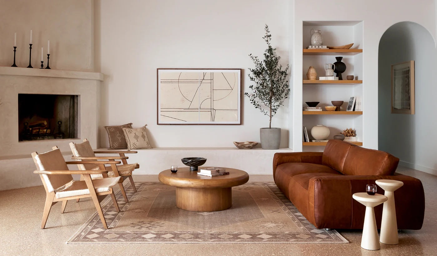 Modern living room featuring a brown leather couch, two wooden chairs, a round coffee table, and a fireplace. A geometric artwork hangs above the mantle, flanked by white walls and built-in shelves displaying art pieces and decor, with a large potted plant nearby.