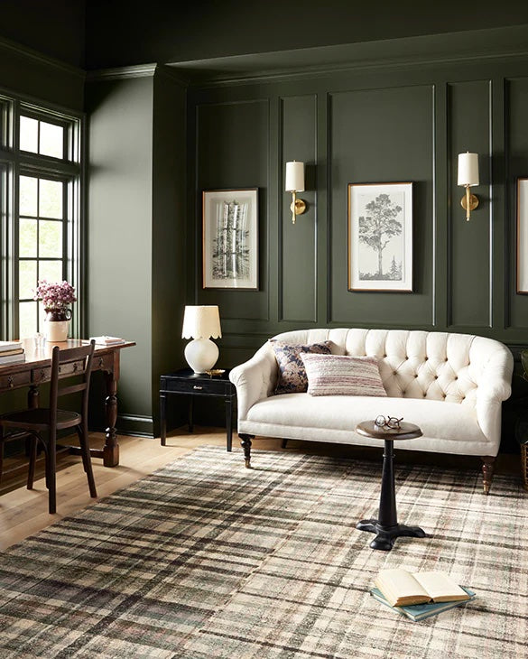 A cozy living room with dark green walls, featuring a white tufted sofa, a small round table, and a plaid rug. Framed artwork and wall-mounted lamps adorn the wall behind the sofa. A desk with a chair and a lamp can be seen by the large windows, allowing natural light in.