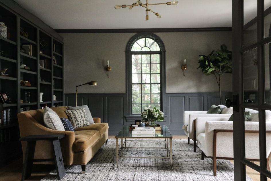 A sophisticated living room with dark gray walls featuring a large arched window. It is furnished with a brown sofa, two white armchairs, a glass coffee table, and shelves filled with books and decor. A potted plant and modern chandelier add elegance to the space.