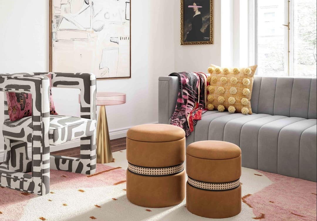 A modern living room features a gray sofa adorned with a textured yellow pillow and a pink throw blanket. Two round brown ottomans sit nearby. Patterned chairs, contemporary artwork, and a window add to the chic, well-lit space. A pink rug covers the floor.