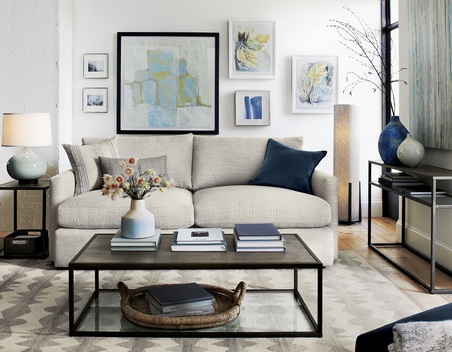 A modern living room with a beige sofa adorned with blue and white cushions. Behind the sofa are abstract paintings. A wooden coffee table with books and a flower vase is in front. There's a lamp on a side table to the left and another lamp on the floor to the right.