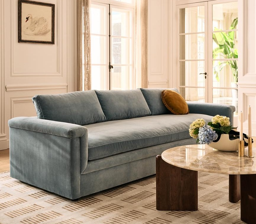 A cozy living room features a light blue, plush sofa with a mustard yellow pillow, positioned against white panel walls. A round marble coffee table with a wooden base holds a vase of blue and yellow hydrangeas next to candles. A framed swan image hangs on the wall.