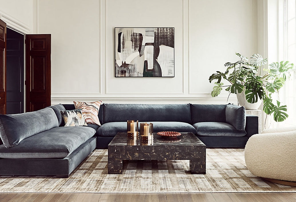 A modern living room with a dark gray sectional sofa adorned with various pillows, a black marble coffee table with candles and decor, a large abstract painting on the wall, and a potted plant by the window adding greenery to the neutral-toned space.