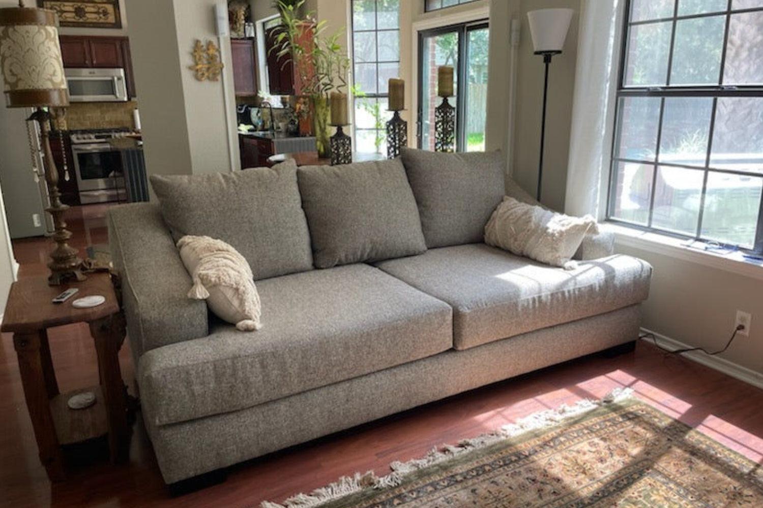 A cozy living room with a gray sofa adorned with pillows, placed by large windows letting in natural light. A wooden side table with lamps and decor items is beside the sofa. A rug is spread over the wooden floor. Kitchen is visible in the background.