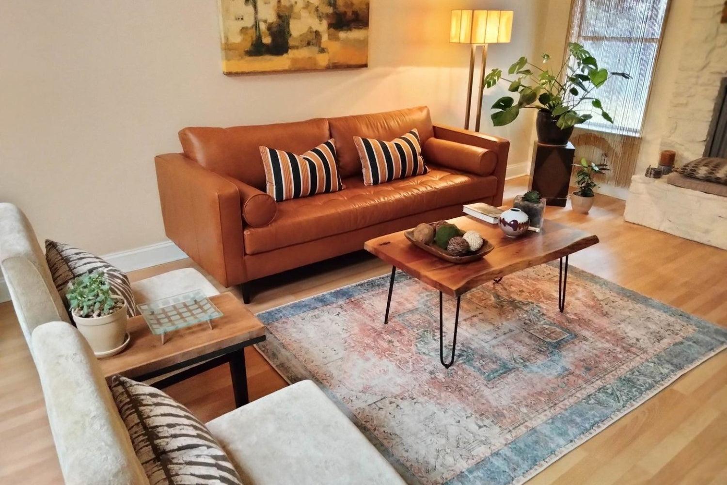 A cozy living room with a brown leather sofa featuring striped pillows, a wooden coffee table with decorative items, a light-colored rug, and two beige chairs. A floor lamp is in the corner near a plant and framed artwork hangs above the sofa.