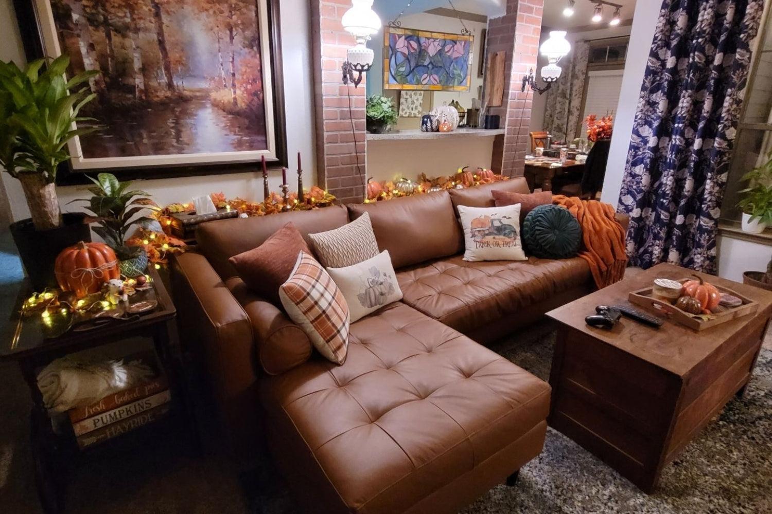 A cozy living room with a brown leather sectional sofa adorned with fall-themed pillows and blankets. Pumpkin decorations and string lights accentuate the autumn ambiance. A wooden chest serves as a coffee table, and leafy plants add a touch of greenery.