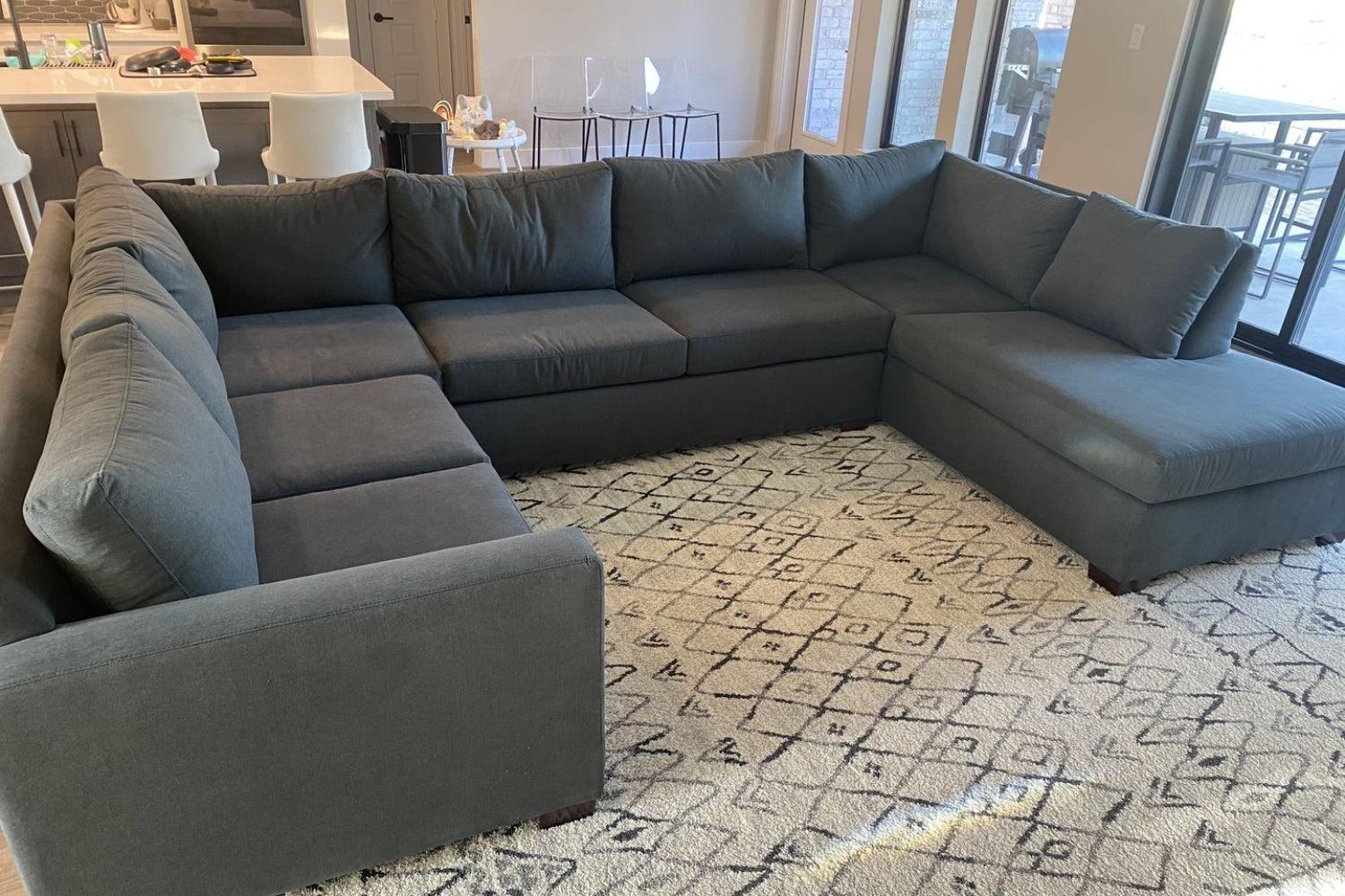 A large, dark gray sectional sofa is set on a patterned rug in a living room. The sofa forms an L-shape, facing a set of sliding glass doors that lead outside. The room features modern decor with a kitchen area visible in the background.