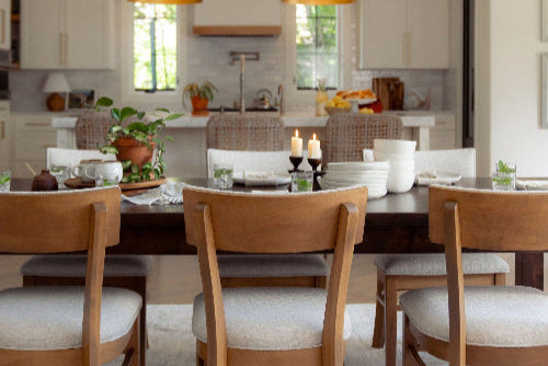 A dining room features a wooden table with six chairs, set with plates and candles. The background includes a modern kitchen with white cabinets, two large hanging lamps, and potted plants for decoration.