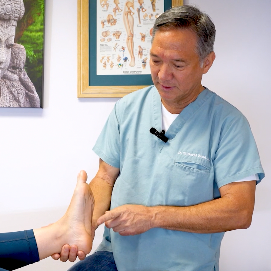 A person in a light blue scrubs top with a name tag examines a patient's foot. The patient‚Äôs lower leg and foot are visible. The practitioner appears to be pointing to a specific area on the foot. The background shows anatomical posters and part of a sculpture.