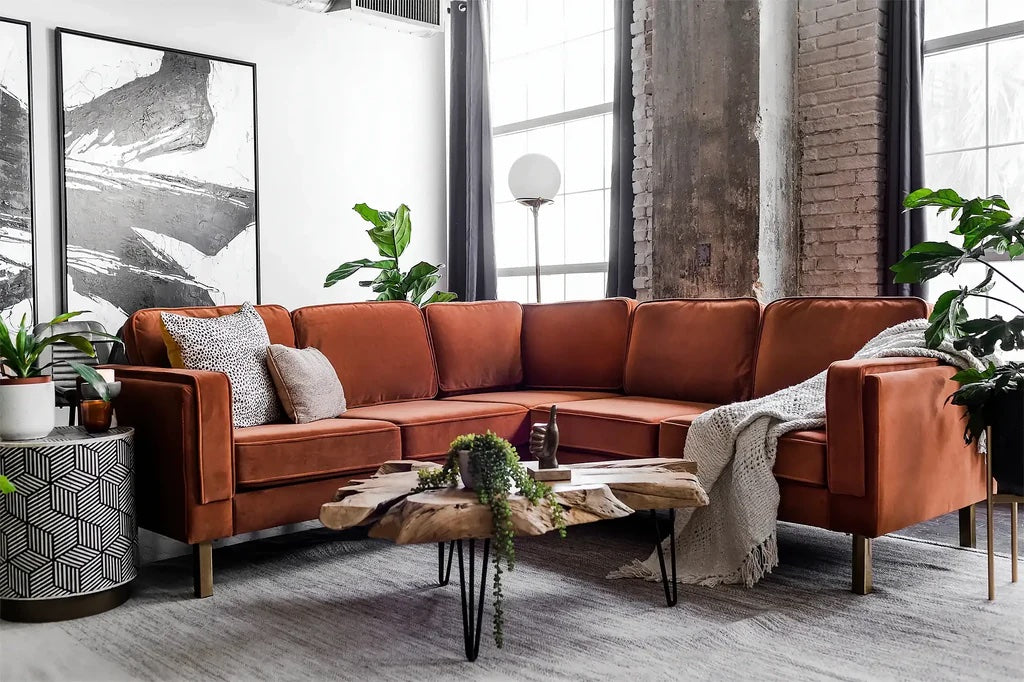 A modern living room features a large, rust-colored sectional sofa with white and gray cushions and a blanket. The rustic coffee table has a natural wood finish and green plants sit atop it. Large black and white wall art and potted plants enhance the stylish space.