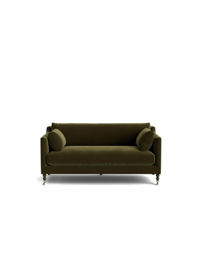 A Fabienne Sofa in green velvet, complete with two cylindrical cushions, set against a plain black background. The sofa boasts a modern design with clean lines and is supported by small casters.