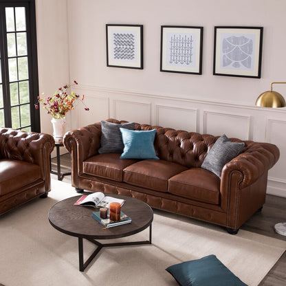 A living room featuring the Birch Lane Freddie 95'' Leather Sofa in brown leather, paired with a matching armchair and accented with gray and blue cushions. A round wooden coffee table displays books and a candle, while three framed abstract art pieces hang on the wall above the sofa.