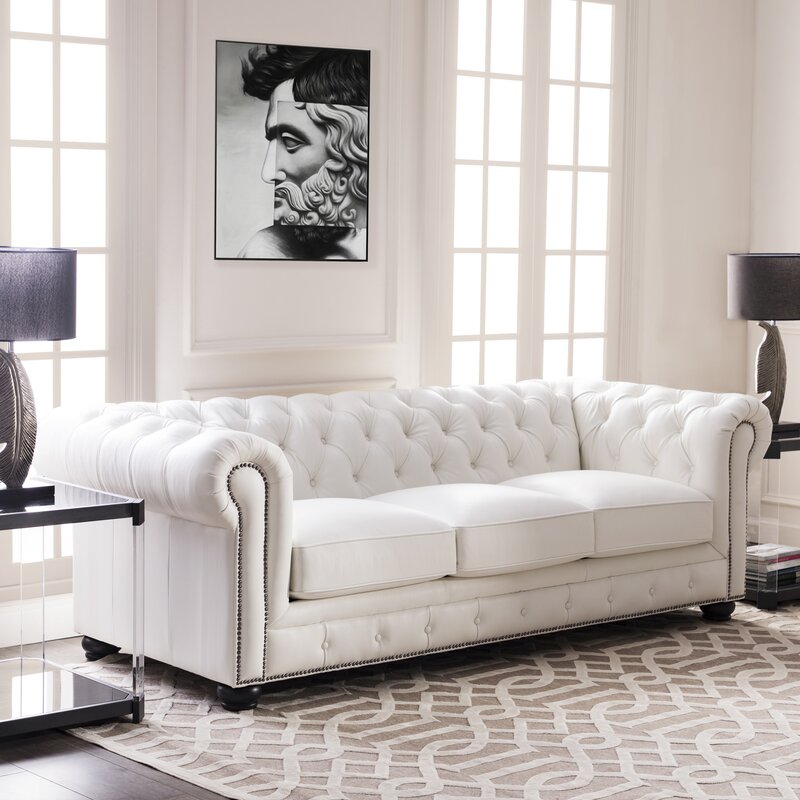 A Birch Lane Freddie 95'' Leather Sofa sits in a bright living room with large windows. A black and white artwork of a face hangs above, while two black lamps flank the sofa, and a patterned rug adds contrast to the floor.