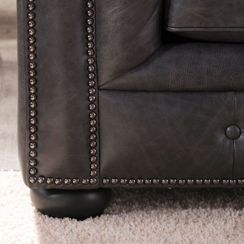 Close-up of the Birch Lane Freddie 95'' Leather Sofa's dark leather corner featuring button-tufted upholstery and metal stud detailing along the edges, placed on a light-colored carpet. The sofa leg is round and black.