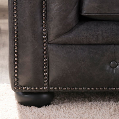 Close-up of the Birch Lane Freddie 95'' Leather Sofa's dark leather corner featuring button-tufted upholstery and metal stud detailing along the edges, placed on a light-colored carpet. The sofa leg is round and black.