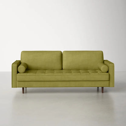 The Geo 84'' Upholstered Sofa, featuring tufted seat cushions, two bolster pillows, and wooden legs, is set against a plain off-white wall on a smooth gray floor.