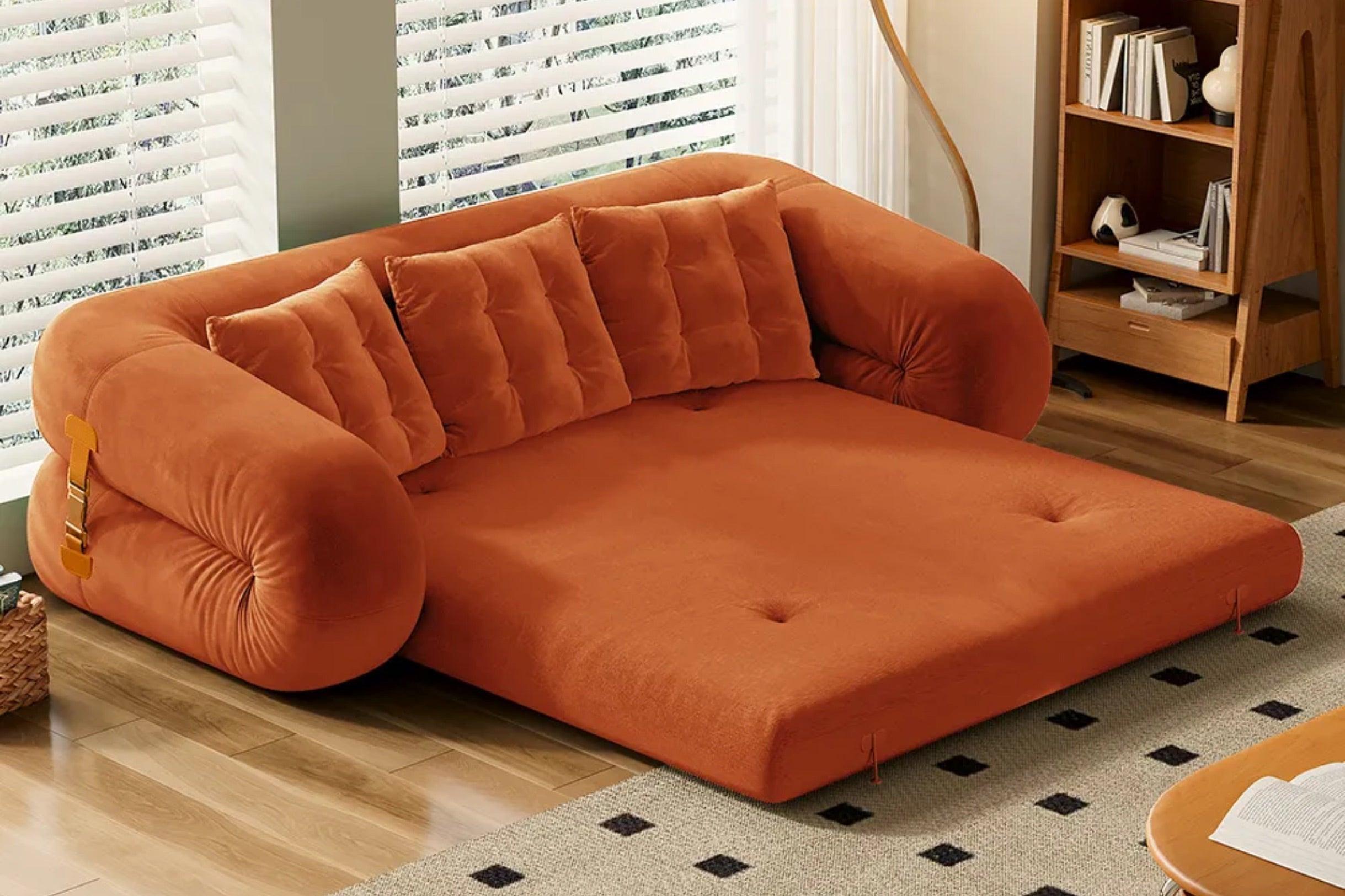 An orange sofa bed is unfolded into a flat position, resembling a mattress. It has several plush cushions against the backrest. The room features a wooden floor, large windows with blinds, and a bookshelf in the background.