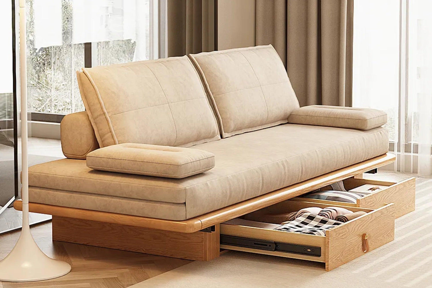 A modern beige sofa with wooden frame and storage compartments underneath. Two drawers are open, revealing a blanket and a laptop. The living room has neutral colors with sheer curtains letting in natural light.