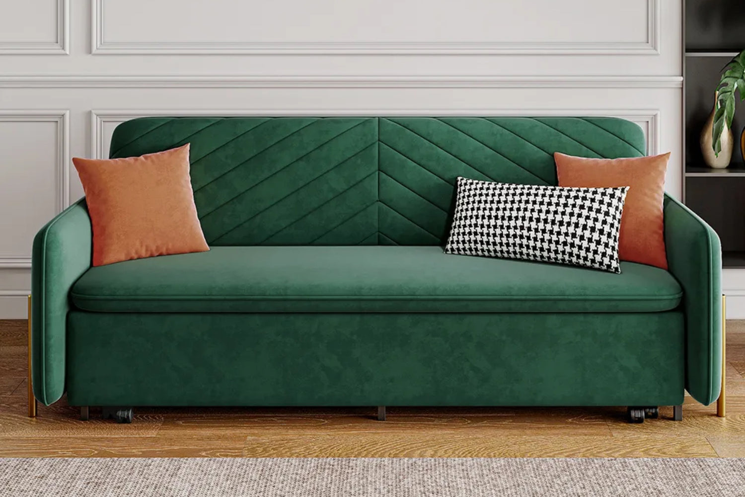 A modern living room features a green velvet sofa with chevron patterns. It is adorned with two orange pillows and a black-and-white houndstooth pillow. The backdrop includes white paneled walls, and there's a small plant on a shelf to the right.