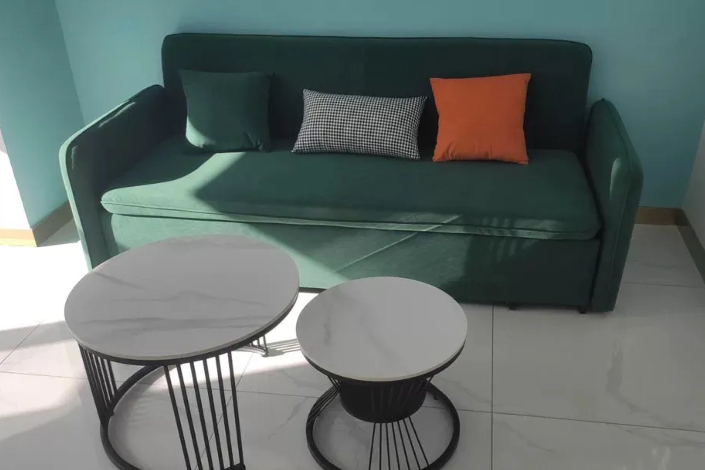 A green sofa with three cushions‚Äîone black, white, and checked; one orange; and one dark green‚Äîagainst a teal wall. In front are two round coffee tables with white tops and black metal bases on a tiled floor.