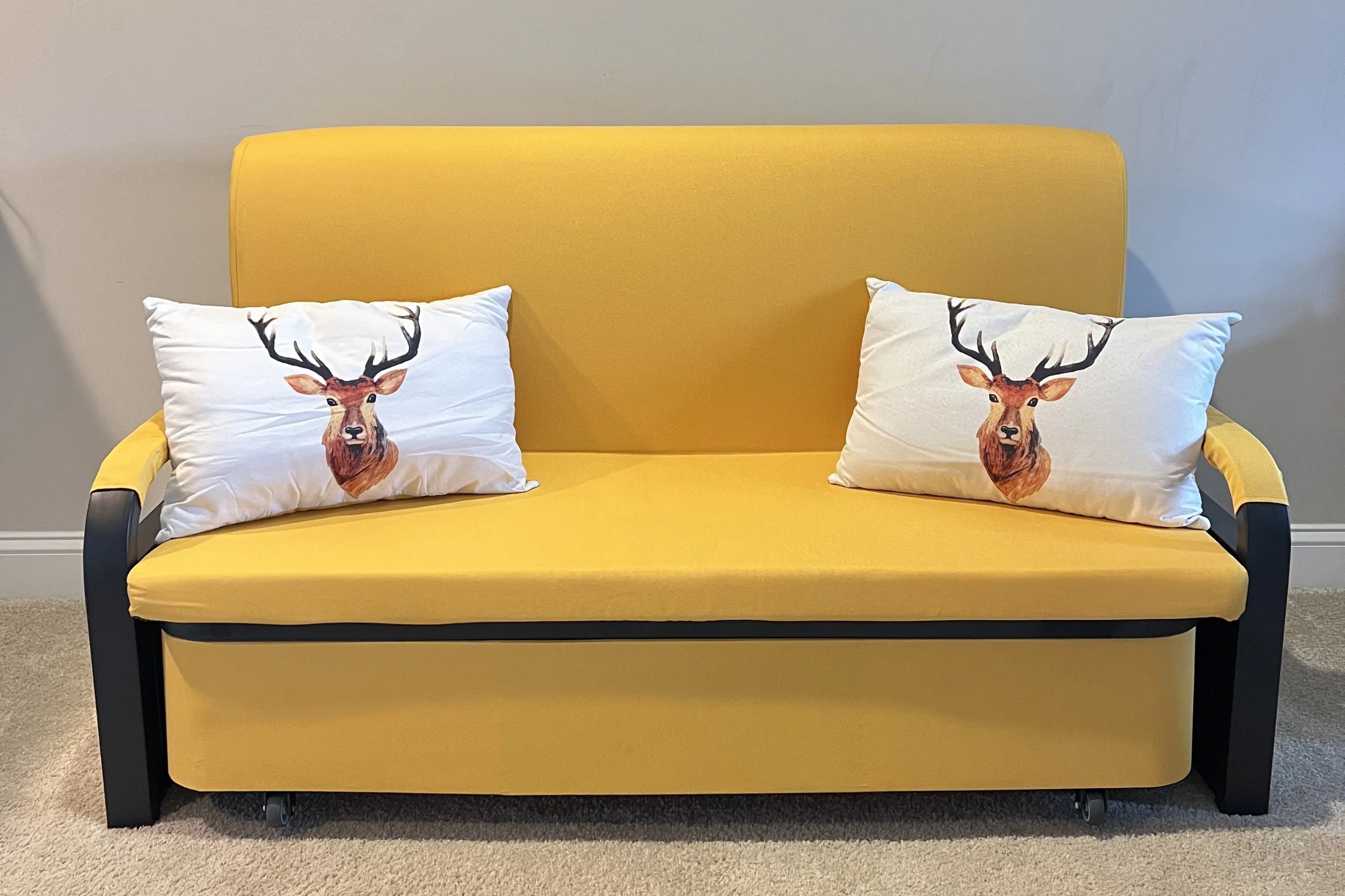 A small yellow sofa with black armrests sits against a neutral wall. Two white cushions with printed images of deer faces are placed on the sofa. The floor is carpeted in light beige.