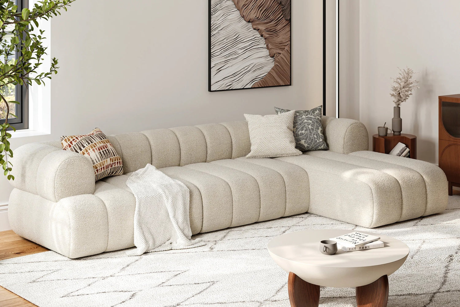 A cozy living room featuring a large beige sectional sofa adorned with patterned pillows and a draped blanket. A round, light wood coffee table sits on a white geometric rug. A nature-inspired artwork and plant add decor elements to the space.