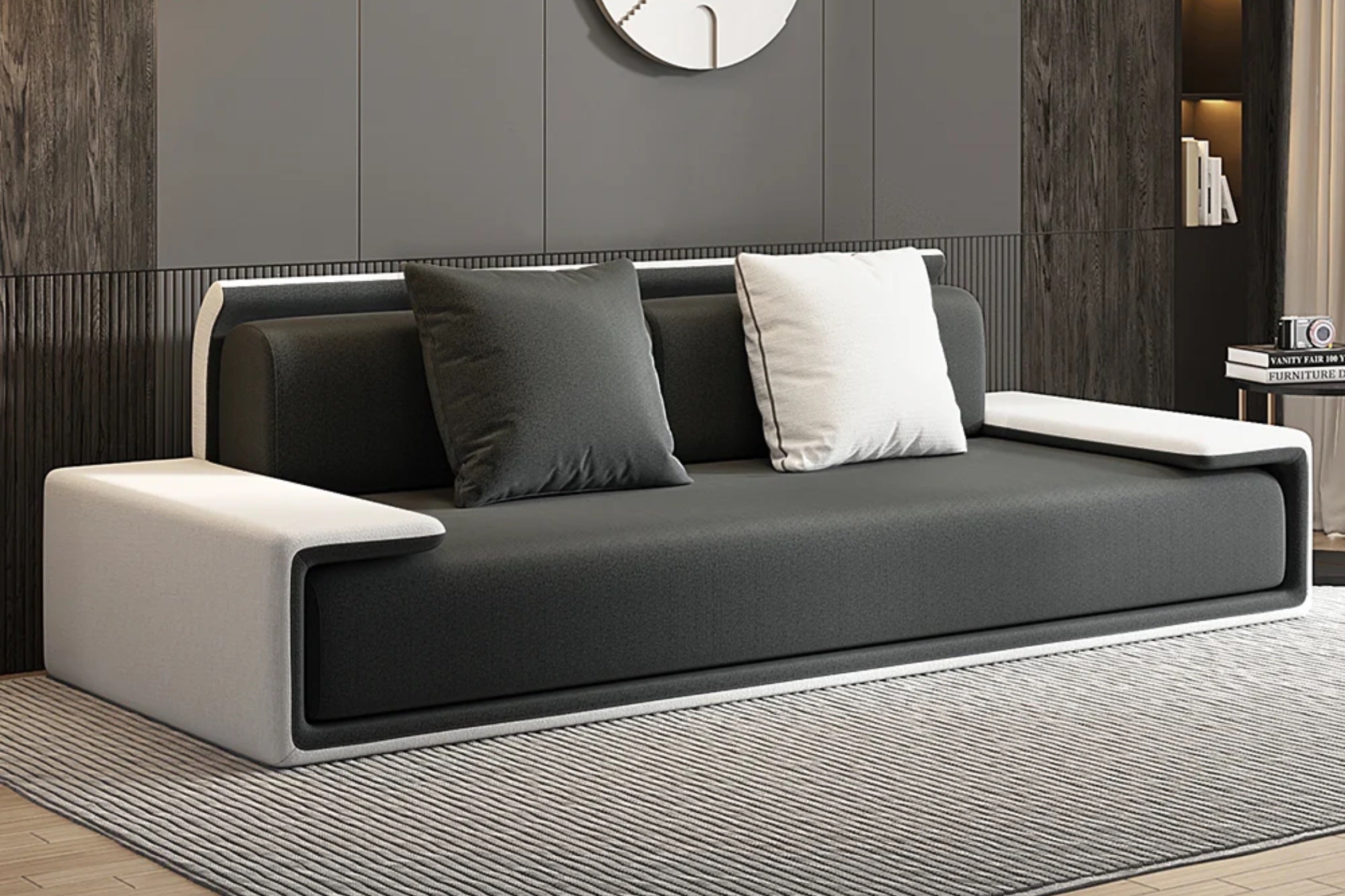 Modern sofa with a sleek black seat and backrest, accented by two black and one white cushions. The arms and base are white, set on a striped gray rug with dark and light wall panels in the background. A circular clock is visible above.