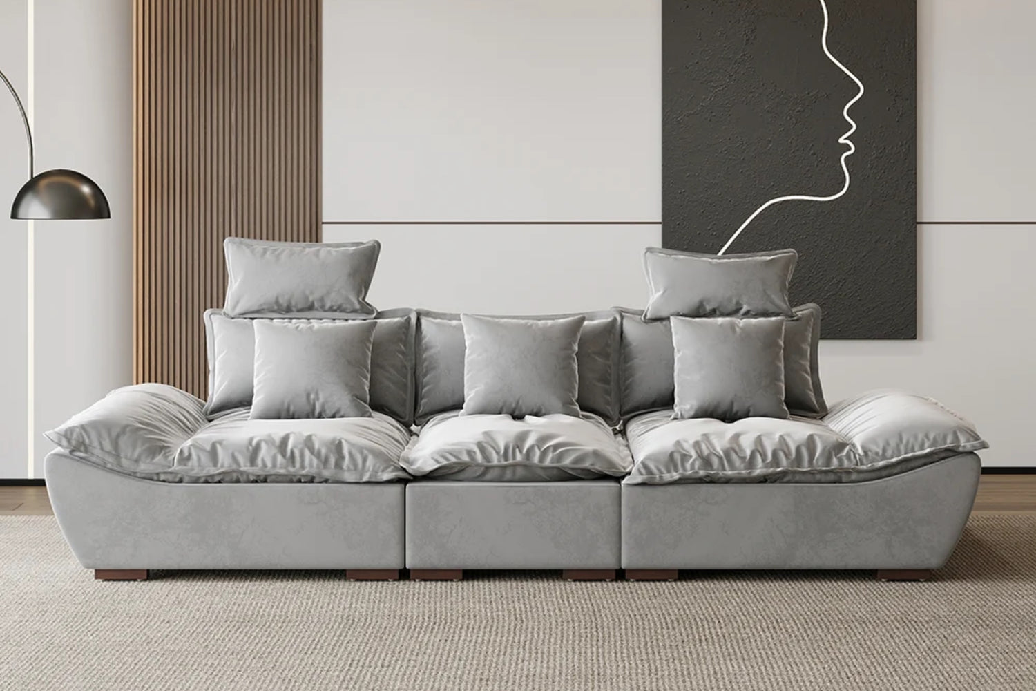 A modern living room with a large, gray, plush sofa and multiple matching cushions. A minimalist black and white artwork of a face profile hangs on the wall. A floor lamp with a dome-shaped shade is on the left. The floor is carpeted.