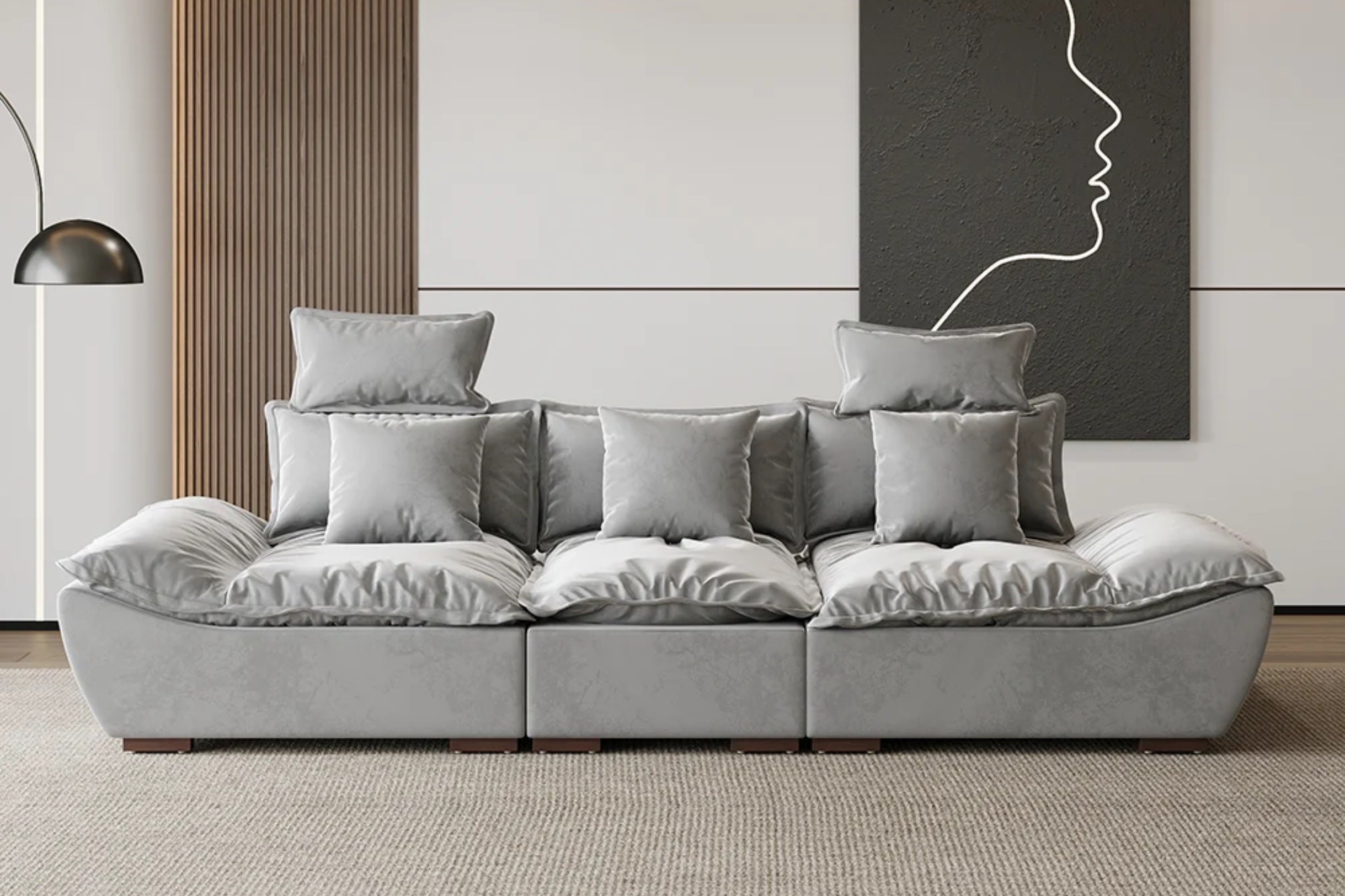 A modern living room with a large, gray, plush sofa and multiple matching cushions. A minimalist black and white artwork of a face profile hangs on the wall. A floor lamp with a dome-shaped shade is on the left. The floor is carpeted.