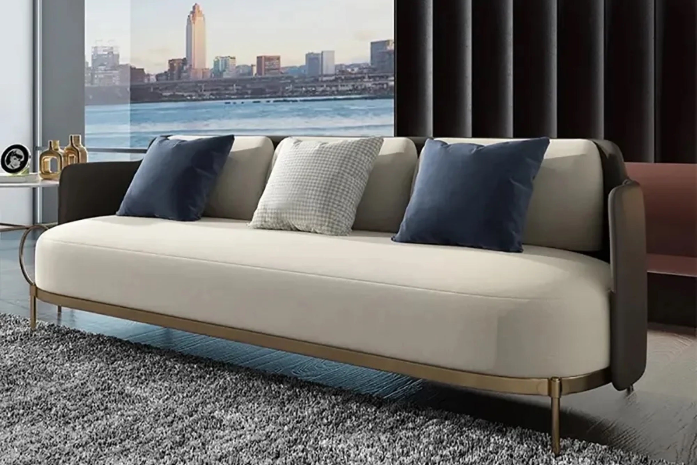 Modern living room with a sleek white sofa featuring two navy blue pillows and a patterned cushion. A large window showcases a cityscape. A textured gray rug is on the wooden floor, with decorative items on a side table.