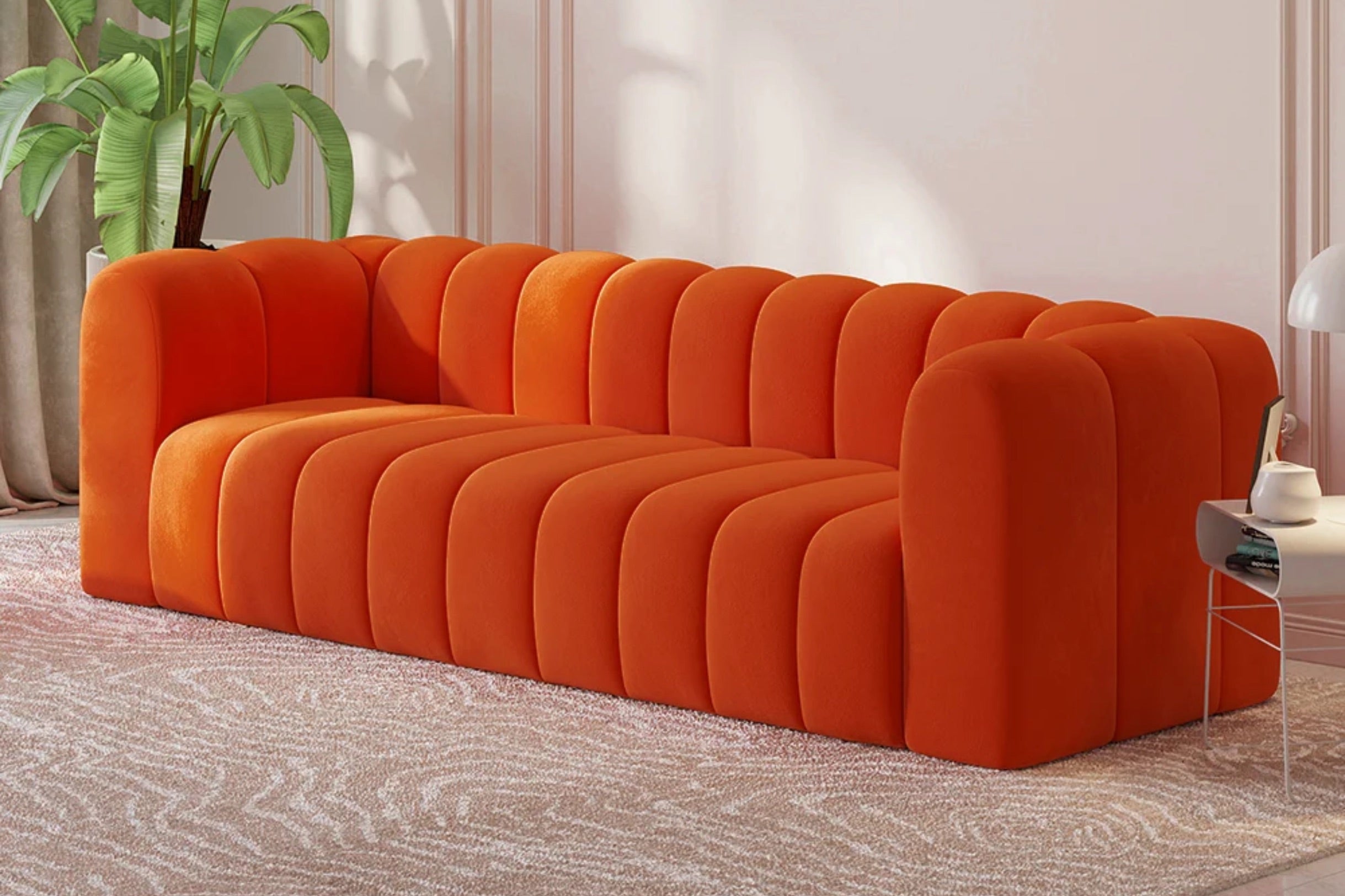 An orange, plush, modern sofa with a ribbed design is placed in a room with light-colored walls and a textured carpet. There is a green plant in the background and a small side table with a white lamp on the right.