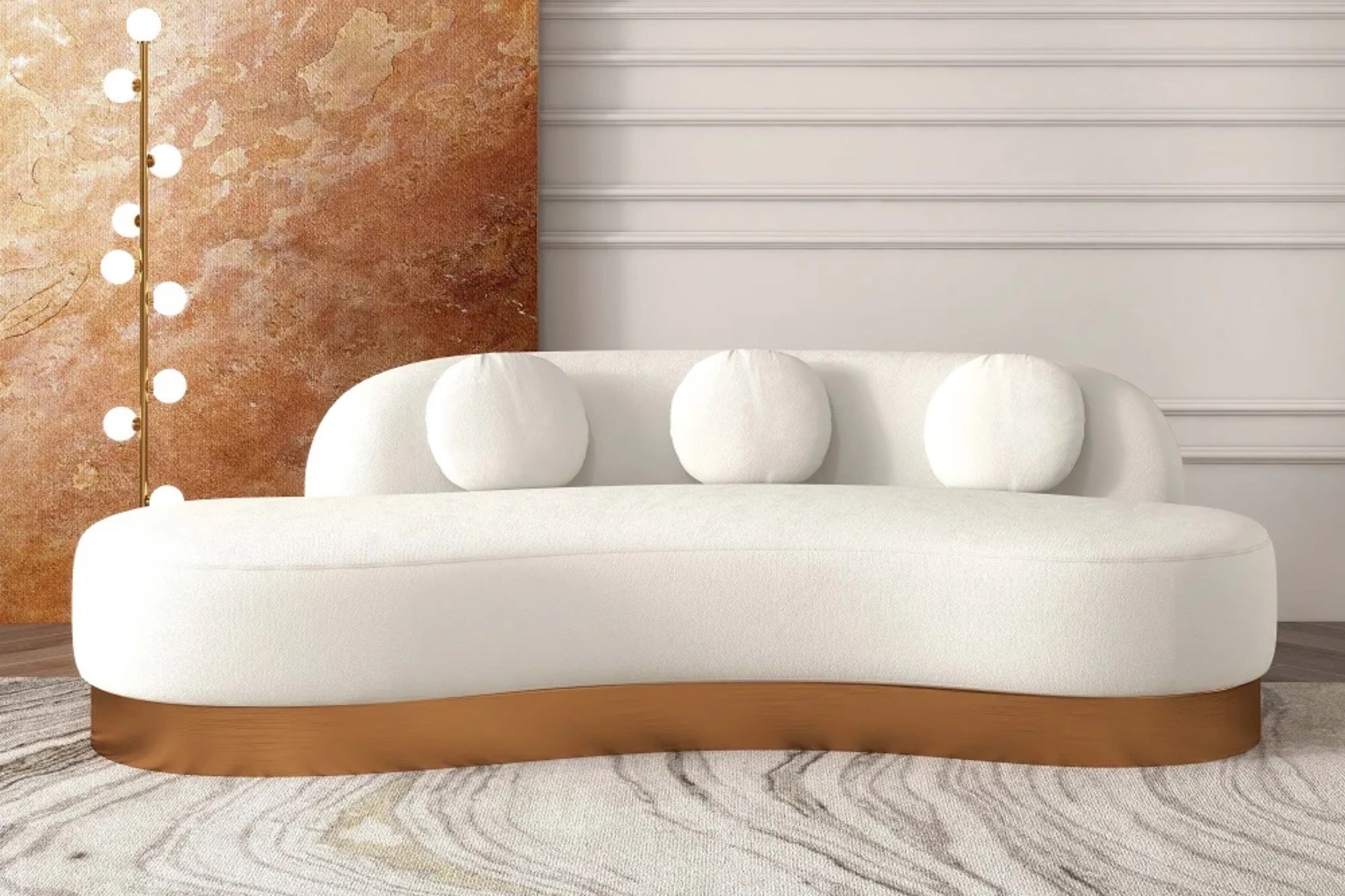 A modern, curved white sofa with a wooden base and three round cushions sits on a textured carpet. A tall floor lamp with exposed round bulbs stands to the side, and a decorative wall with a marbled pattern completes the contemporary setting.