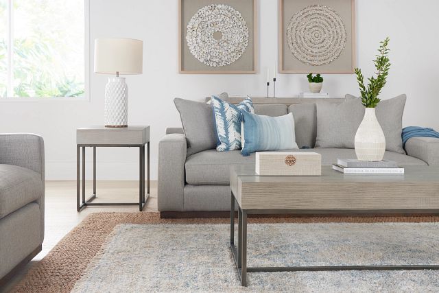 A modern living room featuring a grey sofa adorned with blue and grey cushions, a light grey coffee table with a white vase and book, and a side table with a white lamp. The walls are decorated with two framed circular art pieces. A large window brightens the space.