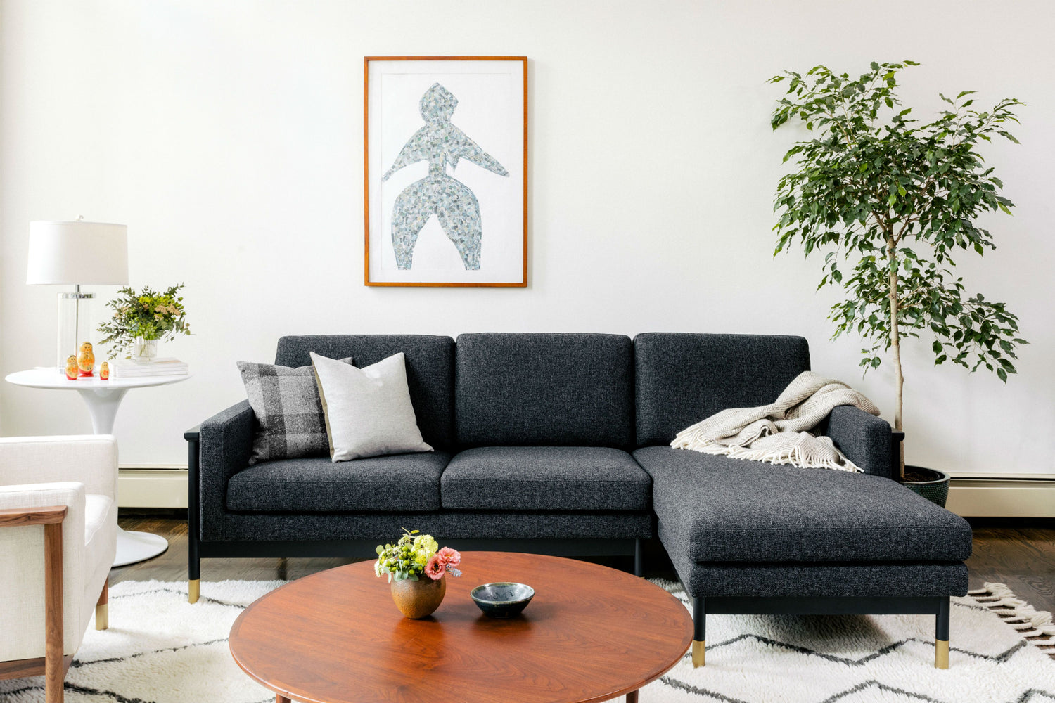 A modern living room features a dark gray sectional sofa with a plaid pillow and a throw blanket. A framed abstract artwork hangs above the sofa. A round wooden coffee table with decorative items sits in front. A large plant and side table with a lamp are in the background.