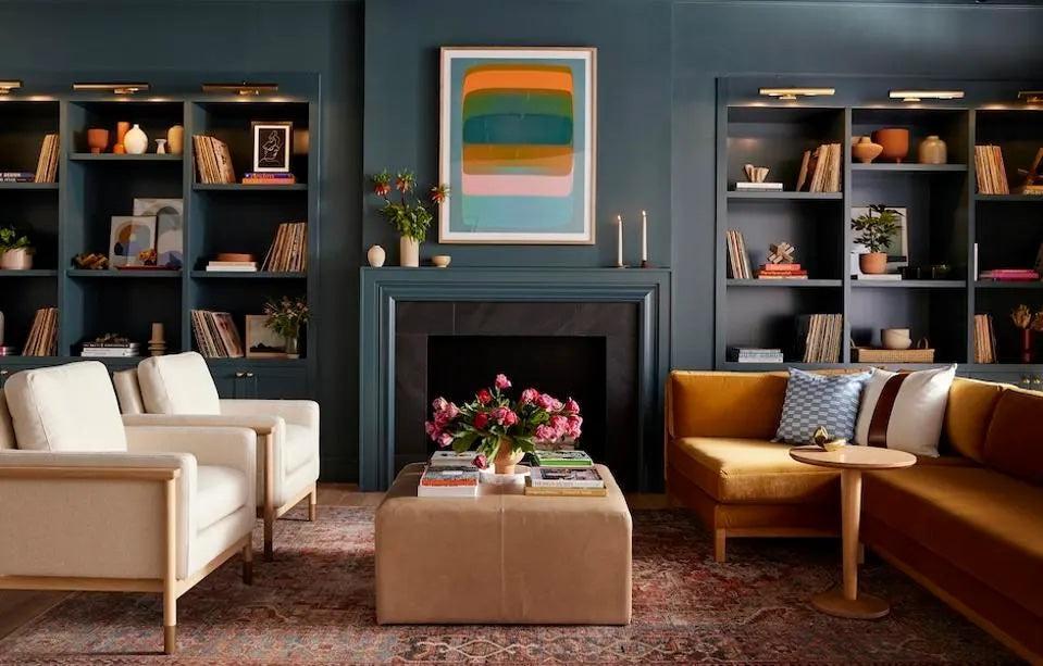 A modern, cozy living room featuring dark blue walls, built-in bookshelves, and a decorative fireplace. The room is furnished with a mustard yellow sofa, two beige armchairs, a central ottoman with flowers, colorful wall art, and a patterned rug.