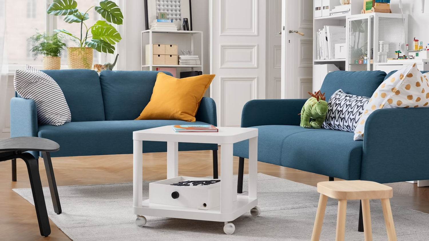 A cozy living room features two blue sofas with various cushions, including a mustard yellow one. A white square coffee table with a book and storage sits atop a gray rug. The room has a wooden floor, a black chair, a small wooden stool, and a potted plant near the window.