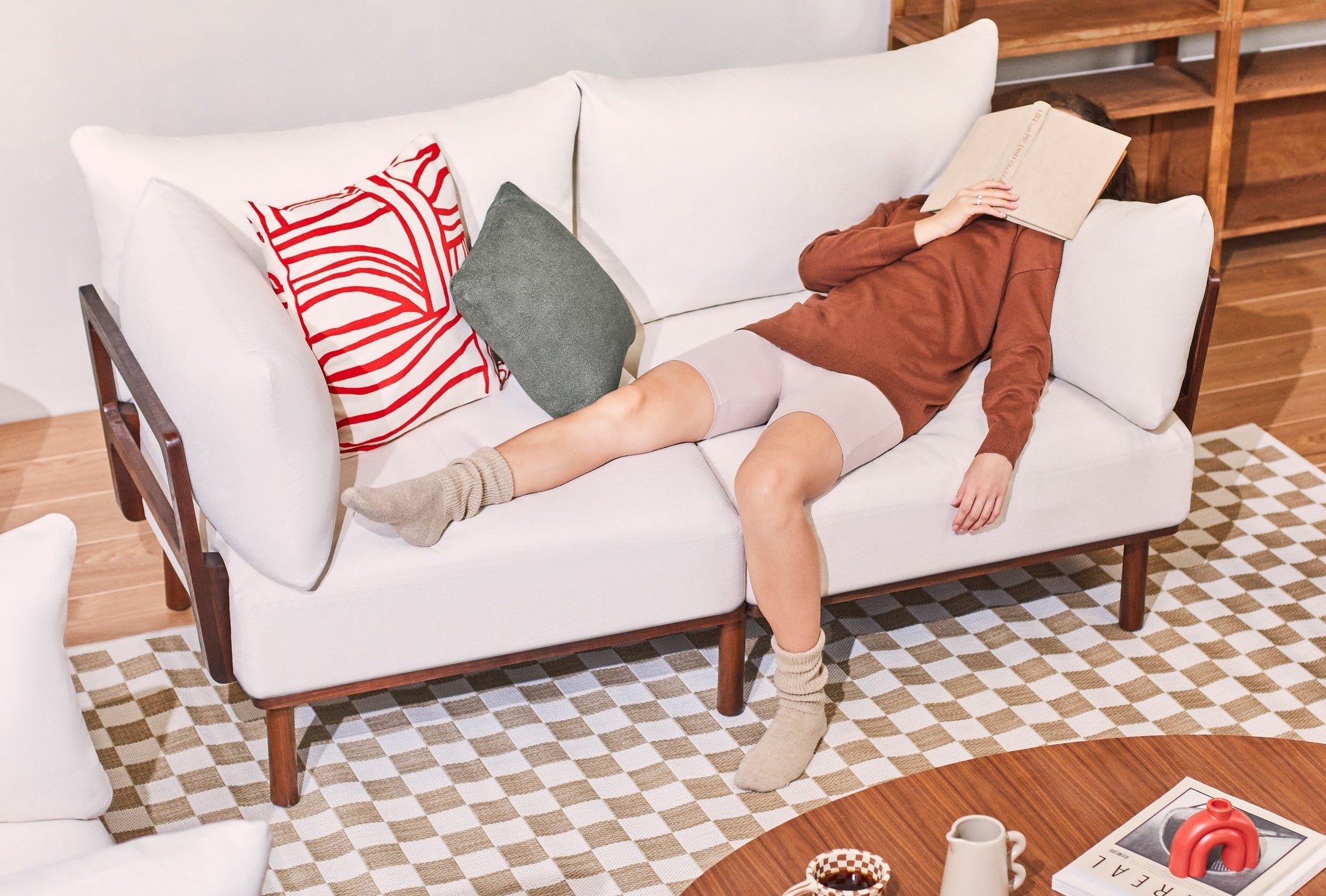 A person is lying on a white sofa with a book covering their face, appearing to be resting or napping. They are wearing a brown long-sleeve top, beige shorts, and beige socks. The sofa has red and green cushions and there is a wooden coffee table in front with various items.
