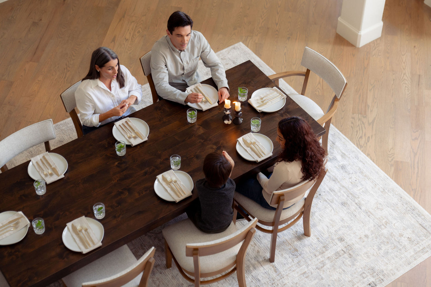 Aerial view of a family of four, sitting around a wooden dining table set for a meal. Each place has a plate, cutlery, and a glass. The room has wooden floors and a light-colored rug. The atmosphere appears warm and inviting.