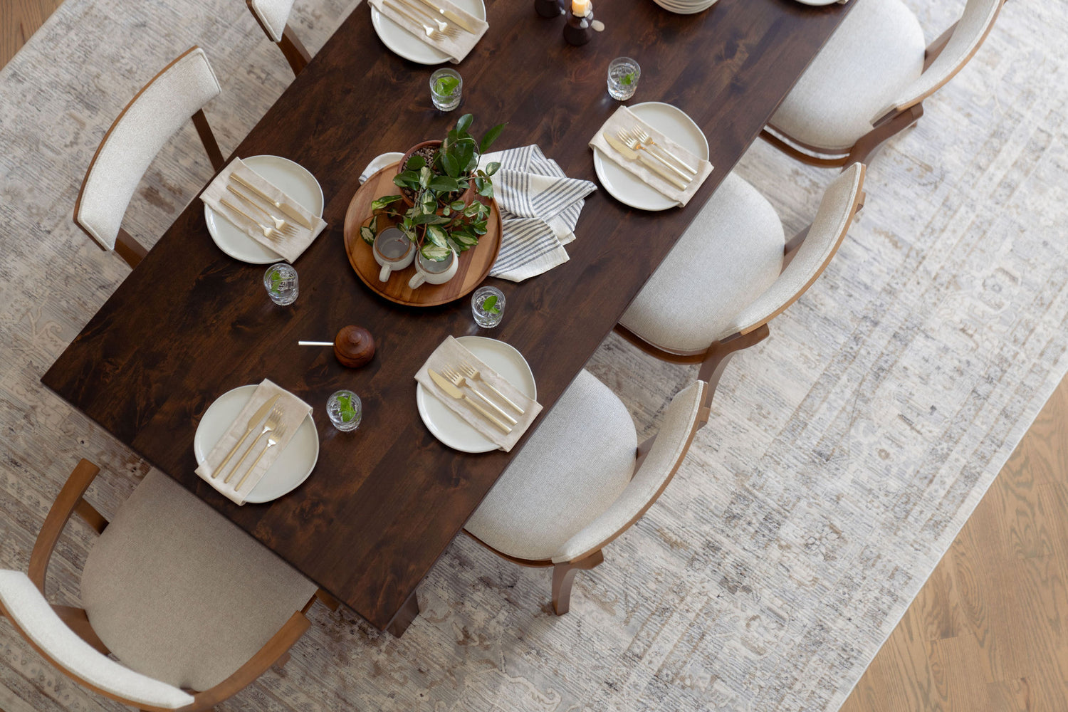 A top-down view of a dining table set for six with wooden chairs. The table has white plates, utensils wrapped in napkins, small drinks, and a centerpiece with green plants. The setting is on a textured area rug.