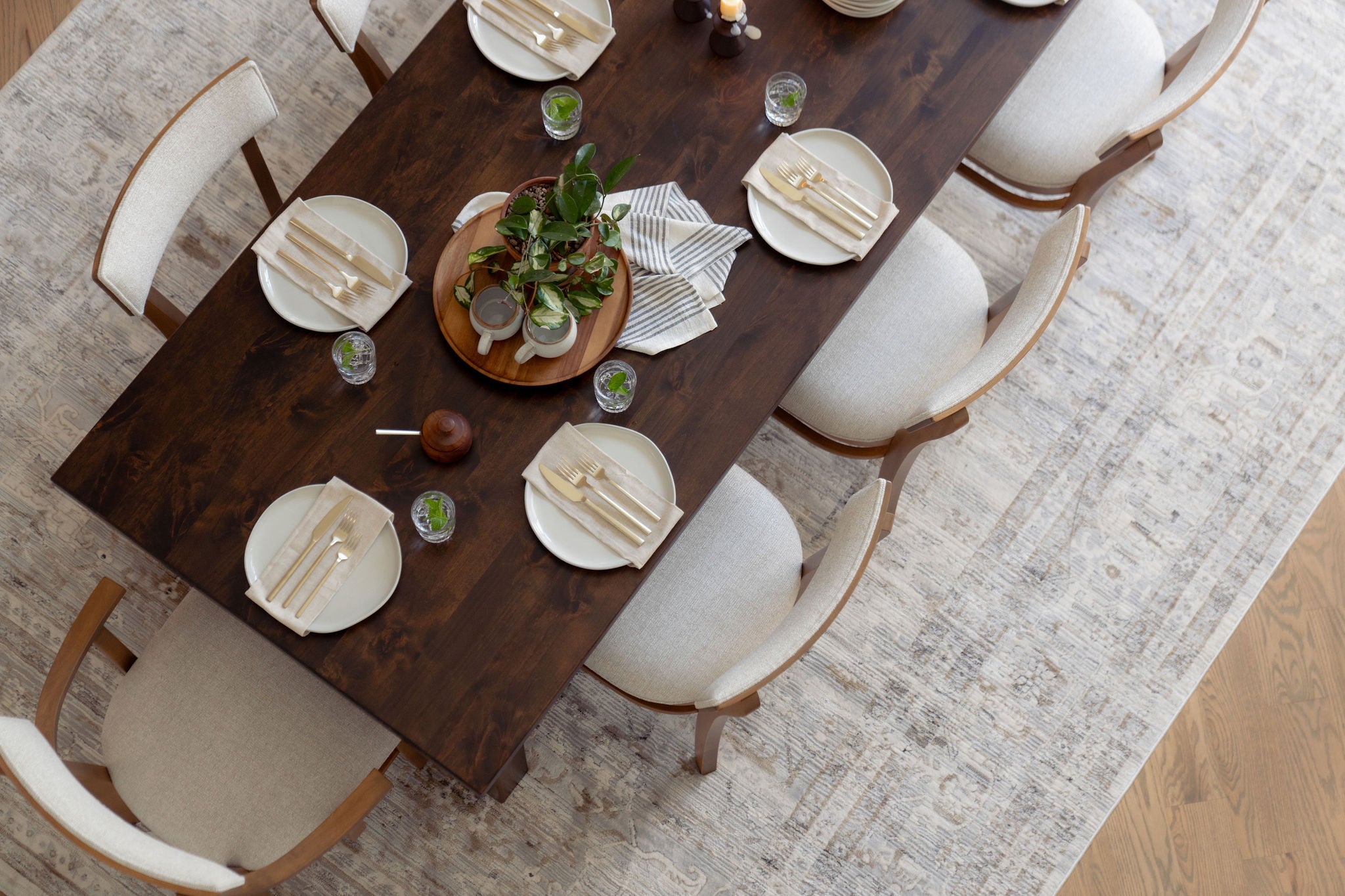 A top-down view of a dining table set for six with wooden chairs. The table has white plates, utensils wrapped in napkins, small drinks, and a centerpiece with green plants. The setting is on a textured area rug.