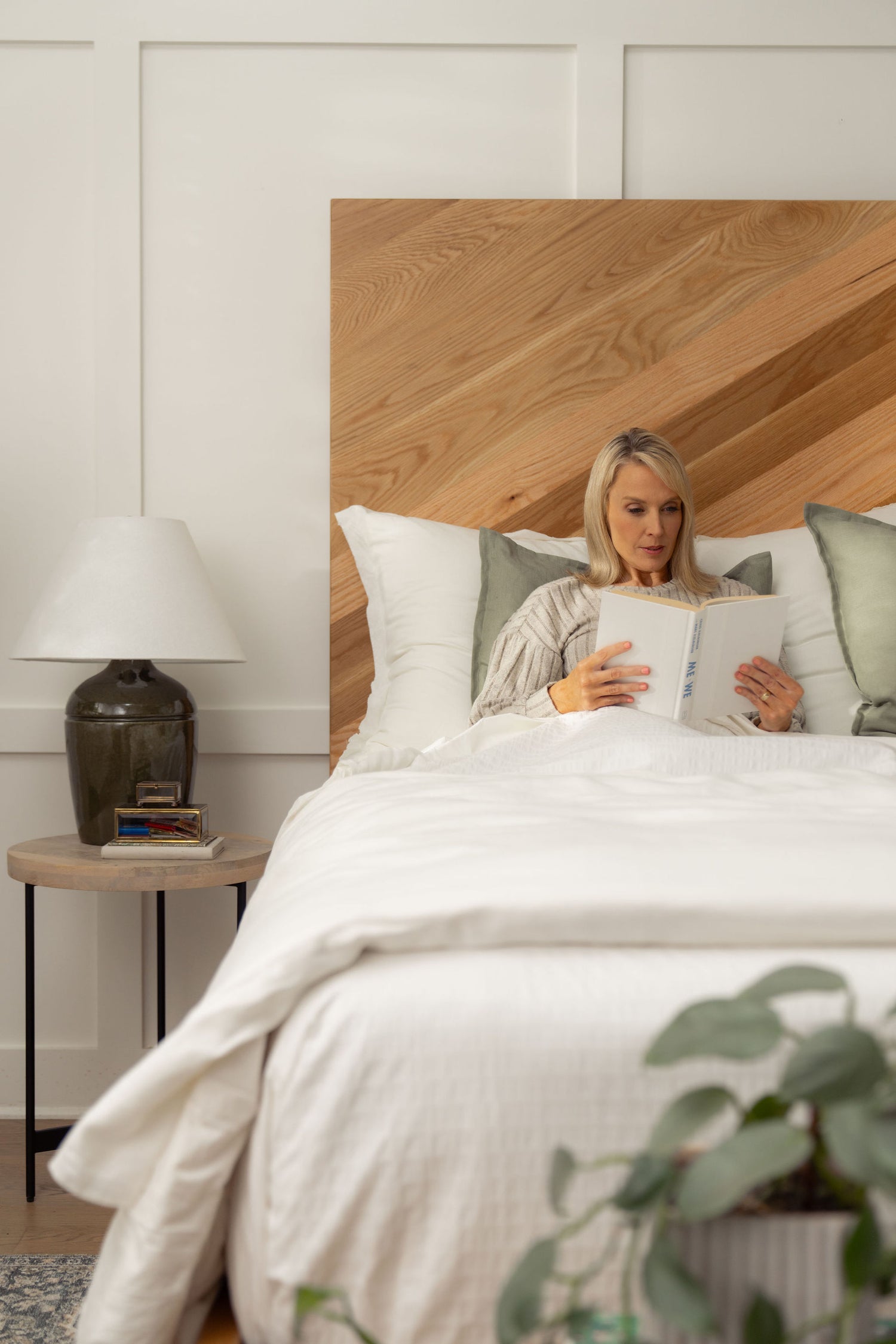 A person with long blonde hair is sitting up in bed, reading a book. The bed has white bedding and green pillows. A wooden headboard and a side table with a lamp and books are visible. There's a plant in the foreground.