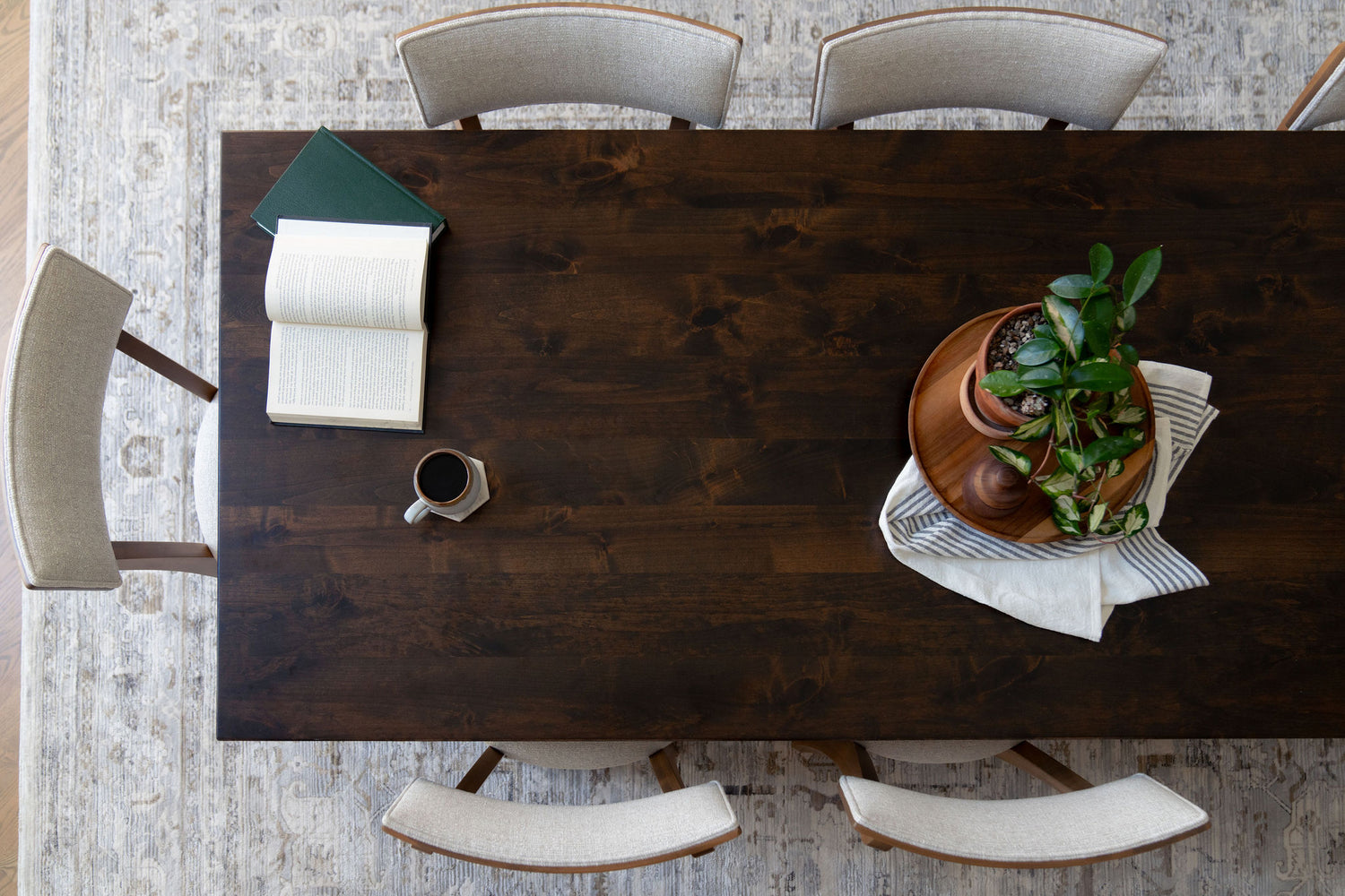 A dark wooden dining table with beige chairs surrounds it. On the table, there is an open book, a closed green book, a cup of coffee, and a centerpiece with stacked wooden trays and a small potted plant.