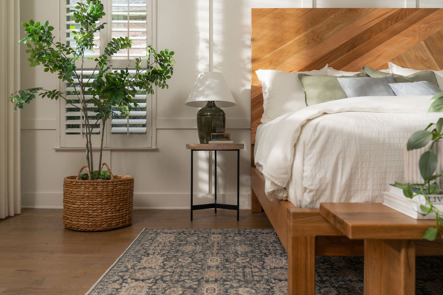 A cozy bedroom featuring a bed with white bedding and green pillows, a wooden headboard, and a side table with a lamp and books. A large potted plant is on the left, next to a window with shutters. A patterned rug is on the wooden floor.