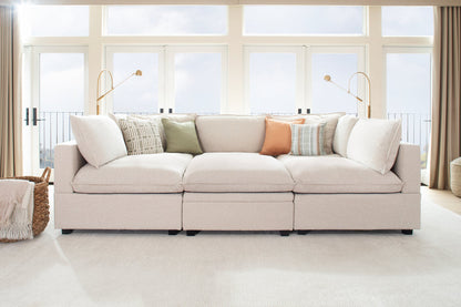 A cozy living room showcasing the Kova Pit, a large beige sectional sofa adorned with patterned and solid-colored throw pillows. Behind the sofa are tall windows with curtains, offering a balcony view, complemented by two modern floor lamps. A woven basket is situated to the left.