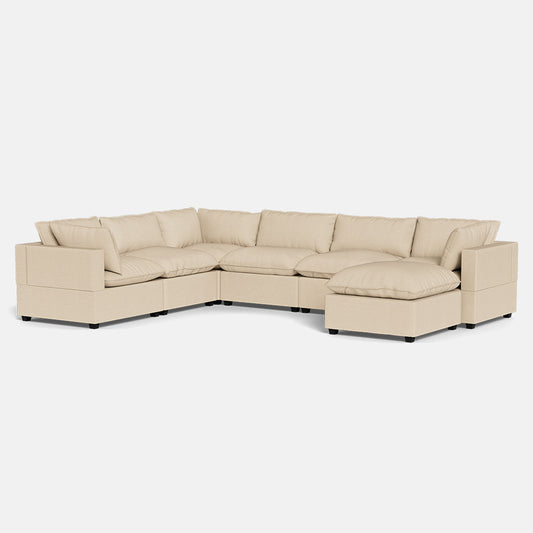 The Kova Pillow Cushion Grand Corner + Ottoman is a large beige L-shaped sectional sofa featuring plush cushions and wide armrests, displayed against a plain white background.