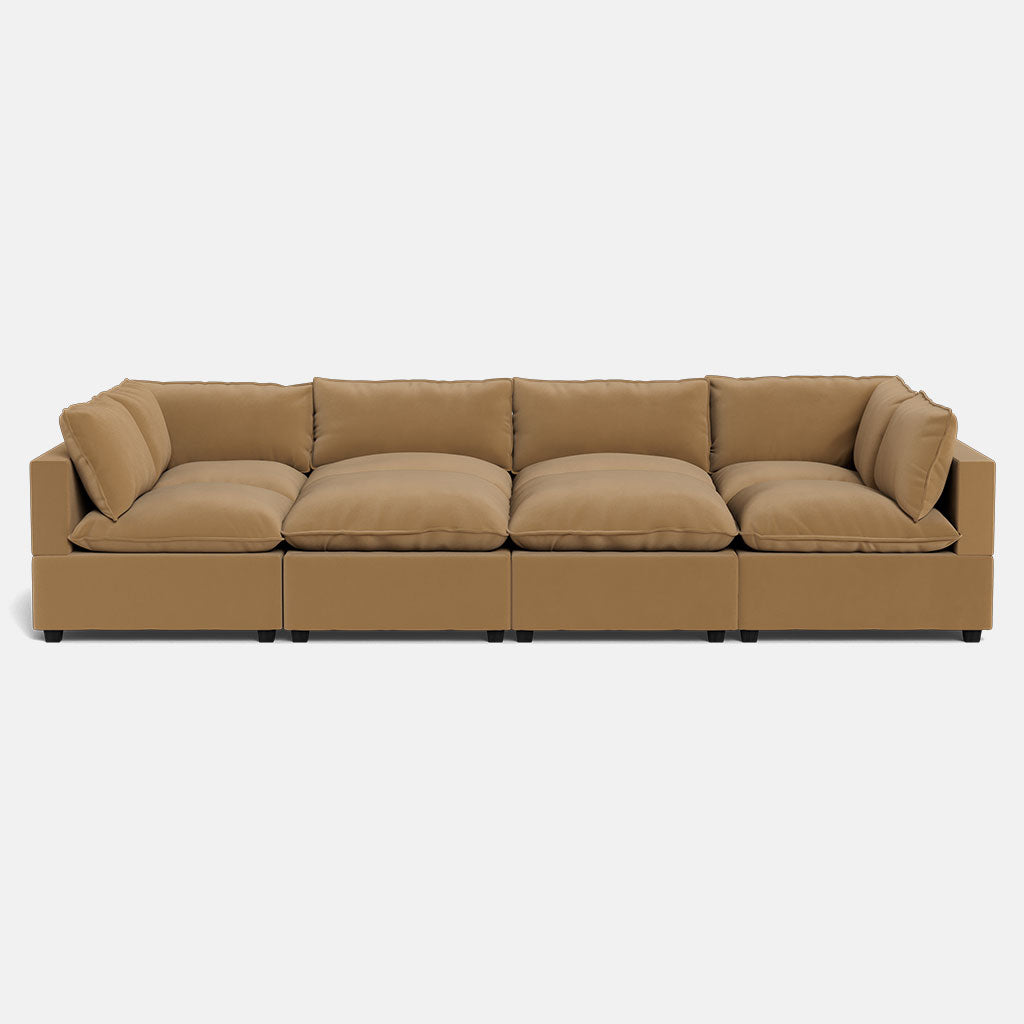 The Kova Pillow Cushion Grand Pit is a modern tan sectional sofa featuring four large seat cushions, plush back and side pillows, and a low rectangular design with short black legs, shown against a plain white background.