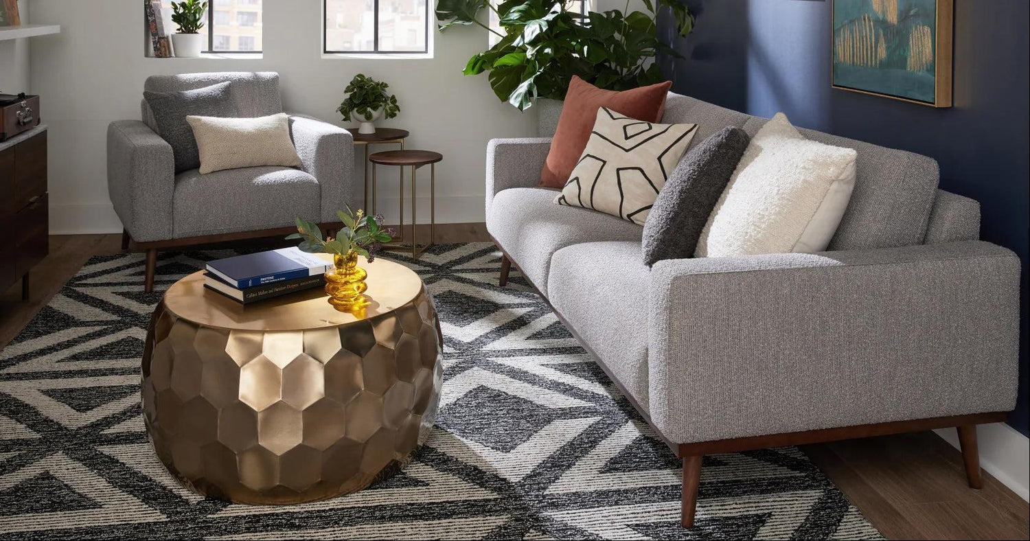 A modern living room featuring a grey sofa adorned with patterned and plain cushions, a matching grey armchair, a hexagonal patterned gold coffee table, and a geometric rug. Indoor plants are near the windows and a painting decorates the dark accent wall.