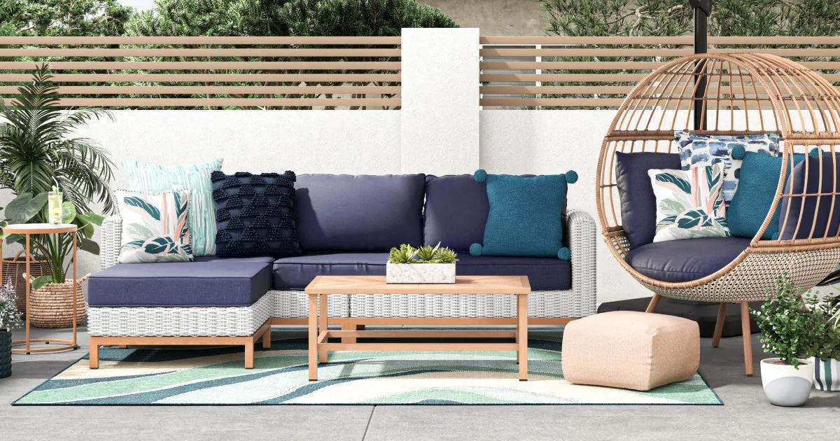 Outdoor patio setup featuring a white wicker sectional sofa with blue cushions and colorful pillows. A wooden coffee table sits in front. To the right, a hanging egg chair with matching cushions. Surrounding are various green plants and an outdoor rug with a wave pattern.